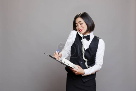 Photo for Smiling asian waitress answering landline phone call and writing on clipboard. Restaurant friendly woman employee in uniform talking with client on telephone and managing order - Royalty Free Image