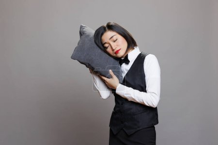 Téléchargez les photos : Fatigué attrayant asiatique serveuse portant uniforme restaurant dormir sur oreiller. Somnolent épuisé jeune femme réceptionniste avec les yeux fermés posant en studio sur fond gris - en image libre de droit