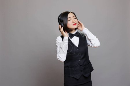 Téléchargez les photos : Serveuse asiatique avec les yeux fermés profitant de la musique dans les écouteurs tout en travaillant. Restaurant travailleuse détendue portant un uniforme professionnel écoutant des chansons dans des écouteurs sans fil - en image libre de droit