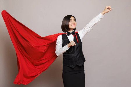 Foto de Sonriente camarera asiática supermujer en velo revoloteando volando, mostrando fuerza y poder. Alegre recepcionista confiado vistiendo capa roja de superman, levantando puño cerrado y mirando hacia arriba - Imagen libre de derechos