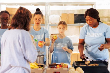Photo for Multiethnic female volunteers sharing fresh fruits and free food to needy, homeless people. African american and caucasian ladies volunteering at food drive, serving the hungry and less fortunate. - Royalty Free Image