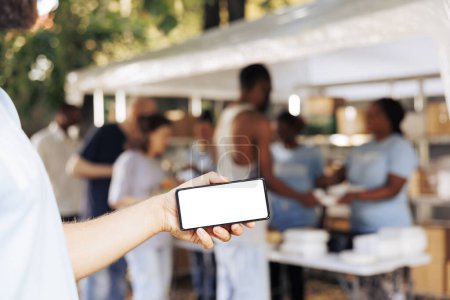 Photo for At charitable food drive, caucasian hand horizontally grasps a mobile phone, displaying an isolated mockup template. Close-up of male volunteer holding a smartphone with blank copyspace white screen. - Royalty Free Image