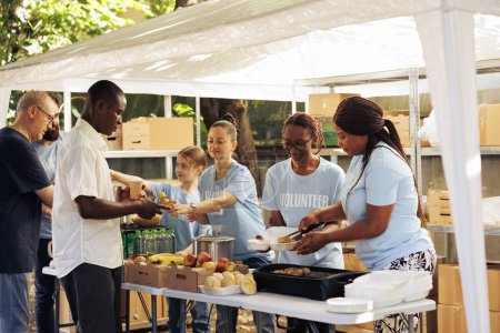 Photo for Individuals of diverse backgrounds gather outdoors to give donated food and non-perishables, providing support to the hungry and homeless. Multiracial volunteers share fresh free meals to poor people. - Royalty Free Image