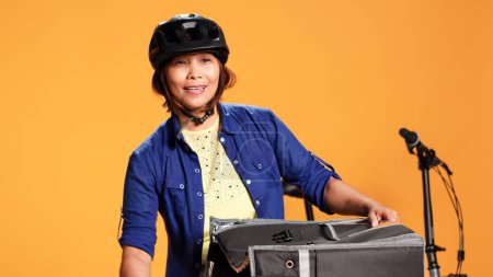 Téléchargez les photos : Joyeux livreur de pizza femme portant un équipement de vélo apportant ordre au client. Gros plan de joyeux coursier BIPOC sortant des boîtes à repas du sac de nourriture thermique, isolé sur fond de studio - en image libre de droit