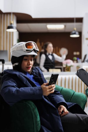 Téléchargez les photos : Pendant les vacances d'hiver, la jeune femme asiatique utilise le téléphone portable dans un coin salon confortable. Femme invitée assise sur un canapé avec un appareil numérique intelligent et des skis de ski portant une veste d'hiver lunettes de ski et casque. - en image libre de droit