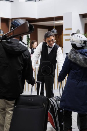 Téléchargez les photos : Couple en vêtements d'hiver arrivant à la réception de l'hôtel et jeune groom asiatique les assiste avec leurs bagages. Employé masculin manipulant les valises des visiteurs à l'arrivée dans la station de ski de montagne. - en image libre de droit