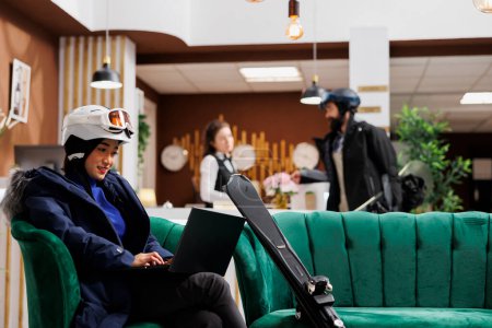 Foto de Mujer asiática con ropa de invierno utiliza el ordenador portátil en la recepción del hotel, mientras que el hombre es asistido por la recepcionista. Invitada femenina con ordenador personal y esquís de esquí sentada en el sofá de la estación de esquí de montaña. - Imagen libre de derechos