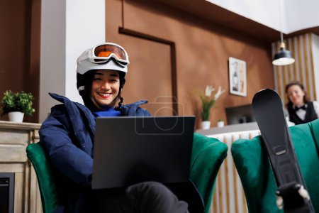 Photo for Asian female tourist wearing winter clothing enjoying her time on sofa with digital device in hotel lobby. Excited woman with skiing equipment browsing on laptop for slopes at ski mountain resort. - Royalty Free Image