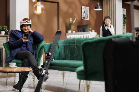 Téléchargez les photos : Femme asiatique enthousiaste utilisant son téléphone portable et attendant dans le hall de l'hôtel de ski pour la procédure de réservation pour les vacances d'hiver. Voyageuse en tenue de sport d'hiver avec son smartphone dans le salon de la station de ski. - en image libre de droit