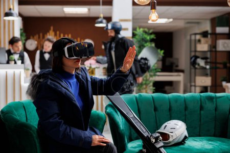 Téléchargez les photos : Femme asiatique avec équipement de ski s'engage avec dispositif de réalité virtuelle dans le salon se préparant pour l'activité de sports d'hiver. Avec des lunettes VR numériques, la station de ski équilibre détente et exploration. - en image libre de droit
