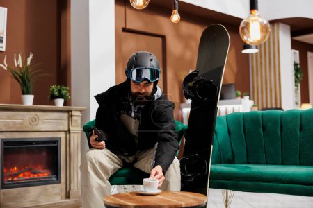 Photo for Tourist with cellphone and snowboard equipment in hotel lobby enjoying beverage. Pictured in winter gear, traveler relaxes near fireplace in ski resort lounge area. Ideal for winter holiday themes. - Royalty Free Image