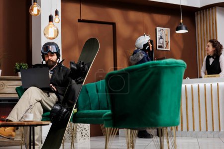 Téléchargez les photos : Homme caucasien portant des vêtements d'hiver assis sur le canapé du hall de l'hôtel et utilisant un ordinateur portable pour la réservation. Le personnel assiste les invités en arrière-plan. Snowboard et lunettes de ski suggère vacances station de ski. - en image libre de droit