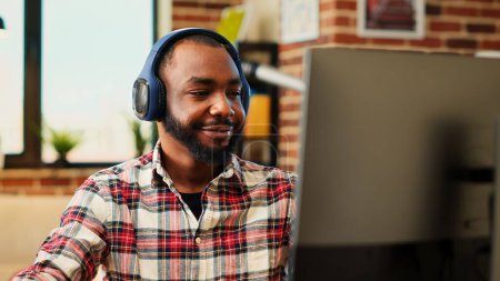 Photo for Happy remote worker listening to music on headphones while working from home. Relaxed smiling freelancer enjoying himself, typing on computer in stylish apartment personal office, close up - Royalty Free Image