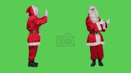 Photo for Santa does no sign and gives thumbs up on both sides on the full body greenscreen studio, wearing festive red suit and hat. Father christmas expressing frustration, doing like agreement symbol. - Royalty Free Image