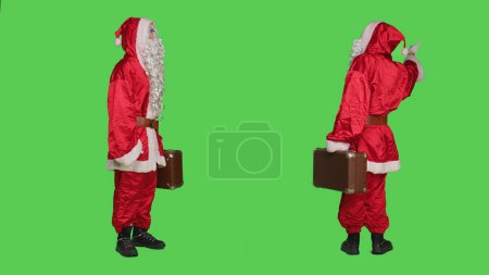 Téléchargez les photos : Le père Noël porte une valise en studio, un costume de fête traditionnel et un chapeau rouge. Caractère Saint Nick avec mallette posant sur fond vert, auto-stop. - en image libre de droit