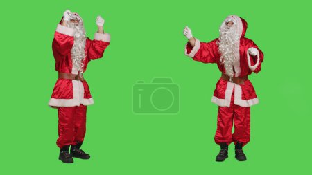Photo for Santa choirmaster conducting band, conductor in winter seasonal costume accompany choir over full body greenscreen backdrop. Character pretends to be musical director, musician in studio. - Royalty Free Image