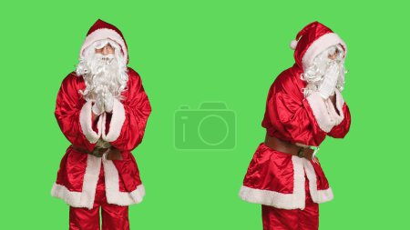 Photo for Saint nick character praying on camera, holding hands in a prayer and posing on greenscreen during holiday season. Young man dressed as santa claus acting spiritual and religious. - Royalty Free Image