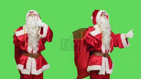 Photo for Father christmas shows thumbs up on camera, feeling positive giving like and agreement symbol over greenscreen backdrop. Santa claus in suit expressing approval with gifts bag. - Royalty Free Image