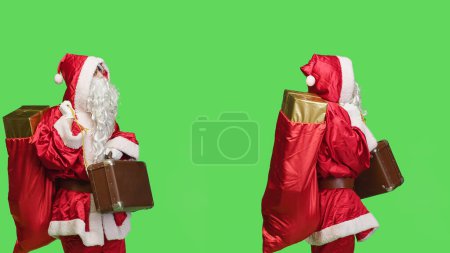 Photo for Joyful adult disguised in santa suit holding suitcase and waiting for something in studio with greenscreen backdrop. Positive person carrying briefcase during holiday season celebration. - Royalty Free Image