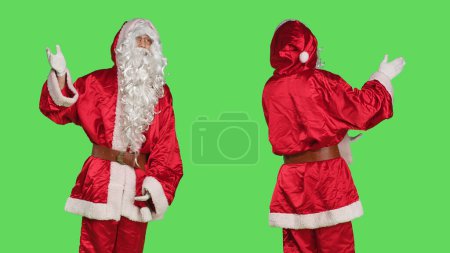 Téléchargez les photos : Santa claus cosplay montre la publicité, pointant de côté alors qu'il pose sur fond vert avec saint pseudo costume de saison. Personne habillée comme père Noël, marketing des vacances d'hiver. - en image libre de droit