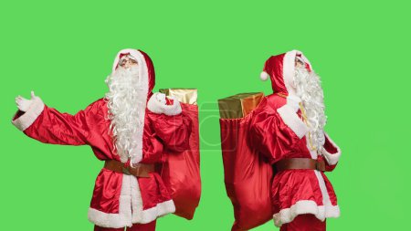 Téléchargez les photos : Homme agissant comme Père Noël claus avec un sac de jouets, portant des cadeaux et des cadeaux sur fond d'écran vert. Saint Nick répandant l'esprit de Noël pendant les vacances d'hiver, sac de transport sur la caméra. - en image libre de droit