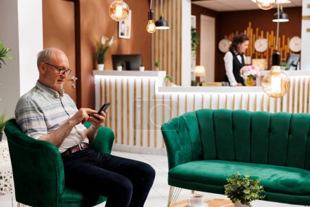 Photo for Elderly male customer seated on sofa using mobile device while waiting for check-in in hotel lobby. Retired senior man surfing the net for holiday activities on his smartphone in lounge area. - Royalty Free Image