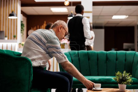 Téléchargez les photos : Au salon élégant pour l'enregistrement, un homme caucasien âgé s'assoit sur un canapé pour prendre une tasse de café sur la table. Homme âgé dans le hall de l'hôtel, attendant sur le canapé pour la procédure de réservation. - en image libre de droit