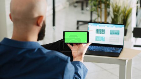 Photo for Worker looking at greenscreen display, preparing to do research for small business plan. Young adult holding smartphone with isolated copyspace mockup, open floor office space. - Royalty Free Image