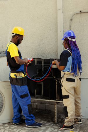 Photo for African american engineers hired after complaints about noisy air conditioner, and warm air coming from supply vents. Technicians using manometers to detect optimum fixing method - Royalty Free Image