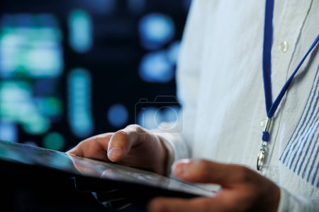 Photo for Engineer in data center using tablet to prevent system overload, close up. Employee in server room ensuring there is enough network connectivity for smooth operations, blurry background - Royalty Free Image