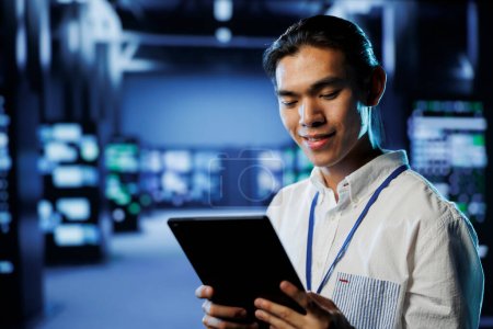 Photo for Cheerful computer operator in data center uses tablet to prevent system overload during peak traffic periods. BIPOC man in server facility ensuring enough network bandwidth for smooth operations - Royalty Free Image