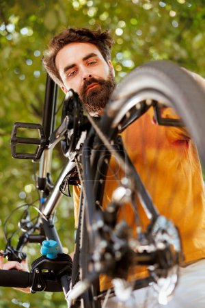 Photo for Male cyclist leisurely checks and maintains his bikes chain in his home yard. Determined young caucasian man examining bicycle parts for damages to repair for outdoor cycling. - Royalty Free Image