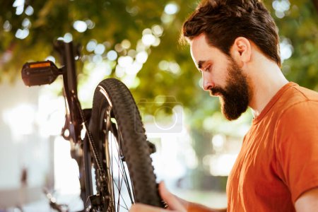 Photo for Outdoor close-up of young caucasian man fastening and changing the rubber on his bike tire for annual maintenance. A determined male cyclist examines damaged bicycle wheel in his backyard. - Royalty Free Image