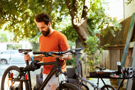 Photo for Athletic caucasian man doing annual maintenance on bicycle while using tools in yard during summer. Young sports-loving male gripping damaged bike for adjustment on repair-stand. - Royalty Free Image