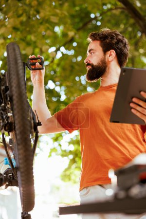 Téléchargez les photos : Cycliste caucasien mâle inspecter et réparer soigneusement vélo avec tablette numérique. Plan détaillé de l'homme sportif en bonne santé recherchant sur la tablette téléphonique pour l'entretien du vélo d'été. - en image libre de droit