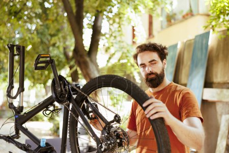 Photo for Outdoor detailed shot of young caucasian man securing and adjusting bike tire rubber for annual maintenance. Determined male cyclist gripping and examining damaged bicycle wheel in home yard. - Royalty Free Image