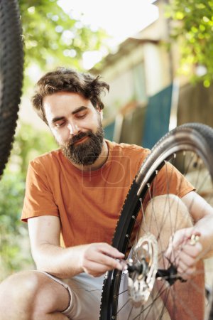 Photo for Healthy sports-loving man maintaining bike wheel for summer leisure cycling. Active young man doing yearly maintenance on bicycle while using tools in the yard during the summer. - Royalty Free Image