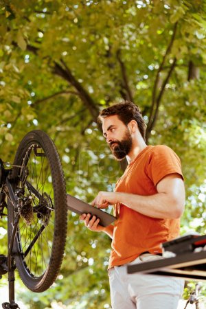Téléchargez les photos : Jeune homme caucasien s'engageant dans l'entretien du vélo fixant habilement roue cassée à l'aide de sa tablette de téléphone. Homme énergique et enthousiaste réparer avec diligence les composants de vélo en utilisant numérique - en image libre de droit