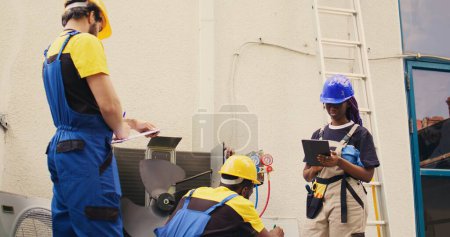 Photo for Trained diverse teamworking servicemen cleaning external condenser internal coolants, checking freon level and calibrating thermostats while coworker checks maintenance procedures on tablet - Royalty Free Image