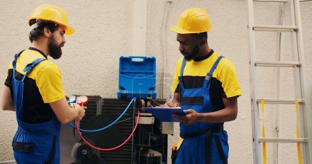 Téléchargez les photos : Expert technicians team working with manifold gauges to check air conditioner refrigerant levels, writing result on clipboard. Skillful mechanics using barometer benchmarking hvac system tool - en image libre de droit
