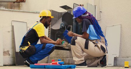 Foto de Trabajadores afroamericanos barriendo escombros sueltos acumulados alrededor de la base del condensador. Ingenieros expertos limpian el cuerpo del sistema hvac y el panel de control con herramienta de cepillo de polvo para eliminar la suciedad suelta - Imagen libre de derechos