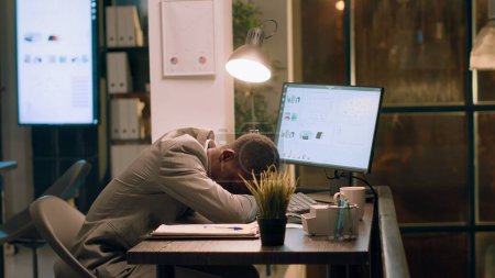 Téléchargez les photos : Homme d'affaires afro-américain surmené endormi dans une chaise de bureau tout en travaillant la nuit au bureau. Employé endormi faisant la sieste pendant le quart de nuit, seul dans l'espace de travail avec des tâches laissées inachevées - en image libre de droit