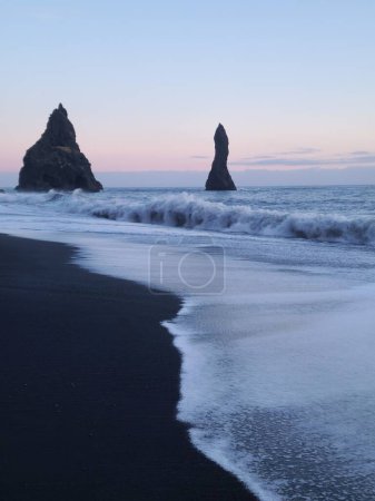 Téléchargez les photos : Reynisfjara plage de sable noir rochers uniques avec ciel rose près de la montagne reynisfjall, belle côte de l'eau. Superbes blocs de basalte sur le rivage formant un paysage naturel icélandique. - en image libre de droit