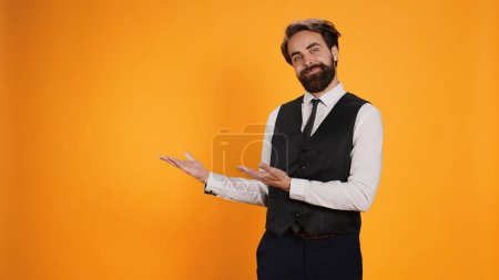 Photo for Skilled classy butler pointing aside and working at five star restaurant. Young professional valet dressed in suit and tie showing direction on left or right sides of yellow studio. - Royalty Free Image