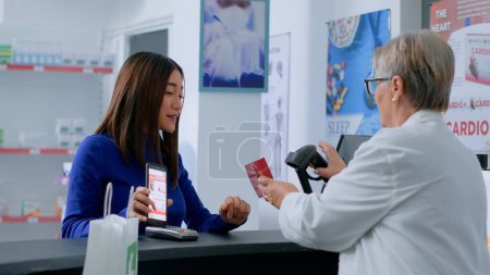 Photo for Customer at drugstore counter, showing druggist prescribed heartbeat regulating pills picture on smartphone, ready to buy it. Aged wellness specialist selling client required treatment product - Royalty Free Image
