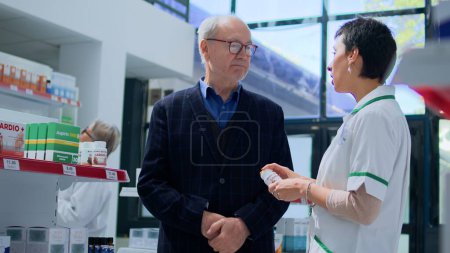 Photo for Aged client in need of prescription sickness treatment drug informations in apothecary, asking experienced pharmacist. Pharmacy supervisor helping elderly customer with pharmaceutical explanations - Royalty Free Image