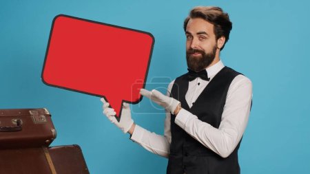 Photo for Hotel concierge shows ad with speech bubble, indicating empty isolated red cardboard icon. Skilled bellboy presents blank copyspace billboard sign in studio against blue background. - Royalty Free Image