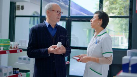 Photo for Aged man in chemist store asking pharmacist for medical opinion, needing treatment for heart ache. Qualified druggist helping senescent client choose best pharmaceutical cure for his disease - Royalty Free Image