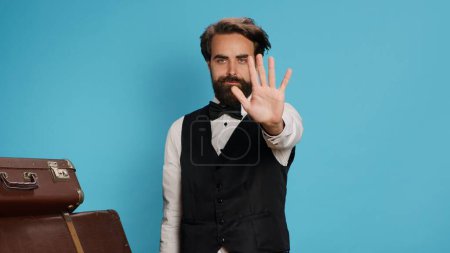 Photo for Doorman raises hand to show stop sign against blue background, portraying advertisement and expressing refusal symbol in studio. Bellboy hotel porter presenting his rejection, luggage bags. - Royalty Free Image