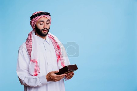 Téléchargez les photos : Homme musulman excité vêtu de vêtements traditionnels tenant boîte cadeau enveloppé avec anticipation. Personne arabe surprise recevant cadeau d'anniversaire, regardant boîte cadeau avec ruban d'arc - en image libre de droit